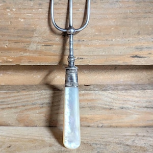 May include: A three-pronged silver fork featuring a smooth, iridescent mother-of-pearl handle. The metal portion includes a decorative, textured collar where the handle meets the tines, showcasing a polished finish and a slender, tapered design.