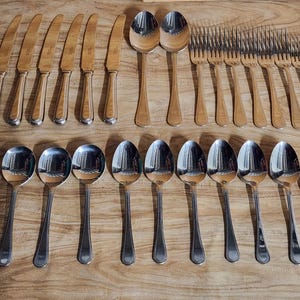 May include: A set of stainless steel cutlery, including knives, spoons, and forks, displayed on a wooden surface. The cutlery has a polished, reflective finish. The knives have gold-toned handles. The spoons and forks are silver.