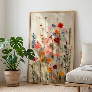 A framed vertical art print displaying a composition of vibrant pressed wildflowers, including red poppies and blue delphiniums, set within a light wood frame. The print is leaning on a wooden floor in a bright, minimalist, Scandinavian-style living room. It is positioned next to a large potted Monstera plant and a light-toned fabric daybed, with a knitted grey rug visible in the foreground, providing a realistic visualization of how the artwork fits a modern, natural interior.