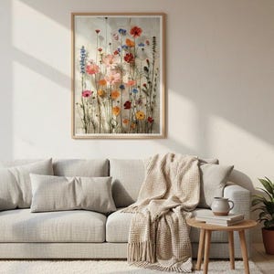 Hochwertiger botanischer Kunstdruck, hängend über einem gemütlichen beigen Sofa im Hygge-Stil. Helles Wohnzimmer mit Strickdecke, Holztisch und warmem Licht. Ästhetische Wandkunst für ein behagliches Zuhause.