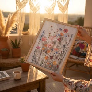 Person holding a framed wildflower art print on a boho-style terrace at sunset. The setting includes warm golden hour lighting, macrame wall hangings, and potted succulents. An aesthetic, lifestyle-focused presentation of botanical wall art for home decor.