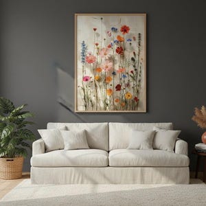 Large 24x36 framed wildflower art print hanging on a dark charcoal accent wall above a bright beige sofa. Boho-style living room decor with a lush monstera plant, pampas grass, and a soft area rug. Stylish botanical wall art for a modern, cozy home interior.