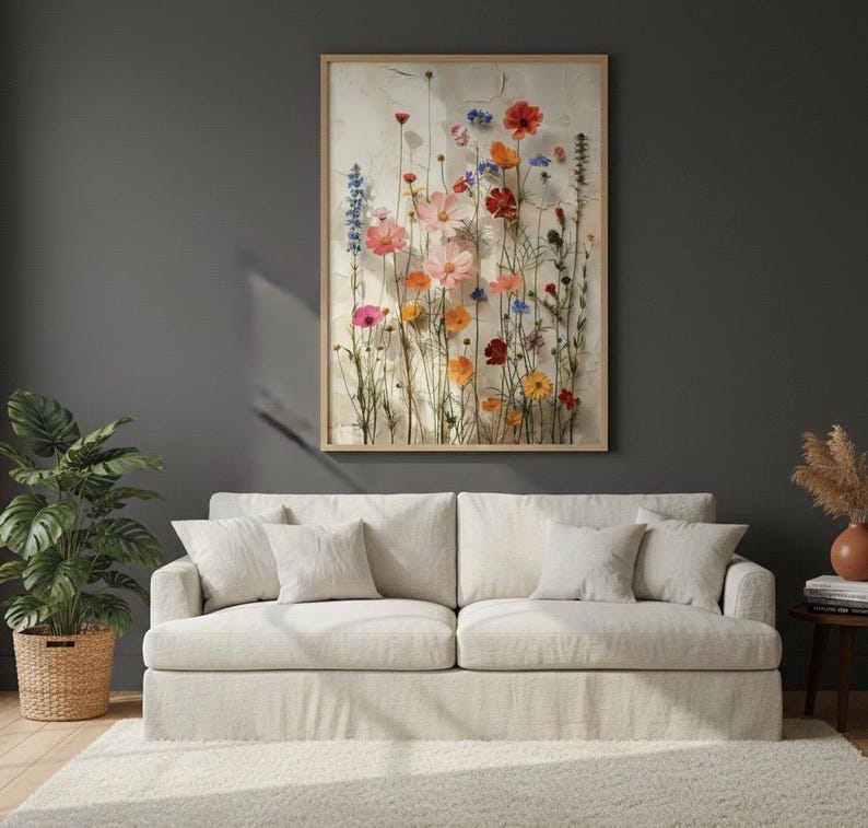 Large 24x36 framed wildflower art print hanging on a dark charcoal accent wall above a bright beige sofa. Boho-style living room decor with a lush monstera plant, pampas grass, and a soft area rug. Stylish botanical wall art for a modern, cozy home interior.