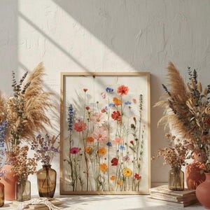 Gerahmter Wildblumen-Kunstdruck, angelehnt an eine Wand in einem künstlerischen Atelier. Umgeben von getrockneten Gräsern in Terrakotta-Vasen und Makramee-Dekoration. Boho-Chic Wohnaccessoire für eine inspirierende, natürliche Inneneinrichtung.