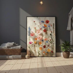Gerahmter Wildblumen-Kunstdruck, angelehnt an eine dunkelgraue Wand in einem gemütlichen Badezimmer. Skandi-Hygge Atmosphäre mit Handtuchstapel, Duftkerze und Pflanze auf hellem Holzboden.