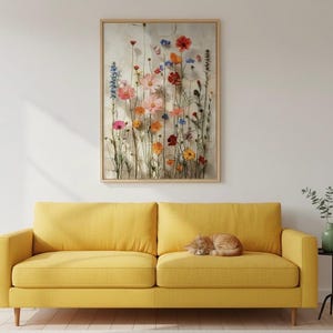 Minimalistisches Wohnzimmer mit einem modernen gelben Sofa, auf dem eine schlafende Katze liegt. Darüber ein gerahmtes Blumenbild mit Wildblumen-Motiv. Harmonische, moderne Wandgestaltung.