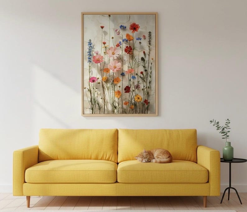 Minimalistisches Wohnzimmer mit einem modernen gelben Sofa, auf dem eine schlafende Katze liegt. Darüber ein gerahmtes Blumenbild mit Wildblumen-Motiv. Harmonische, moderne Wandgestaltung.