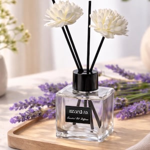 Op de afbeelding: Een heldere, vierkante etherische olieverspreider met een zwarte dop en rietjes, bekroond met twee witte bloemvormige decoraties. De diffuser staat op een houten dienblad met takjes paarse lavendel. Het label luidt "Essential Oil Diffuser."