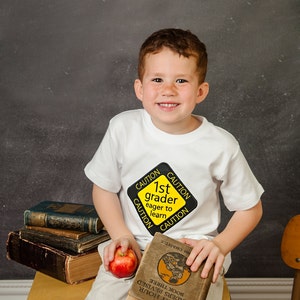Może przedstawiać: Młody chłopiec w białej koszulce z ż&oacute;łtym znakiem ostrzegawczym, na kt&oacute;rym widnieje napis "1st grader eager to learn", siedzi na drewnianym krześle trzymając książkę i jabłko.