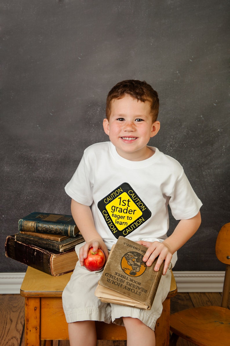 Może przedstawiać: Młody chłopiec w białej koszulce z ż&oacute;łtym znakiem ostrzegawczym, na kt&oacute;rym widnieje napis "1st grader eager to learn", siedzi na drewnianym krześle trzymając książkę i jabłko.
