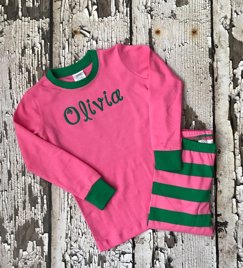 May include: Pink and green children's pajamas with long sleeves and a green stripe pattern on the pants. The top has the name "Olivia" embroidered on the front in green.
