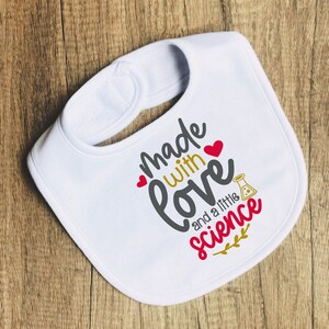 May include: White baby bib with the text "Made with love and a little science" in black and red lettering. A small red heart and a beaker with a red liquid are included in the design.