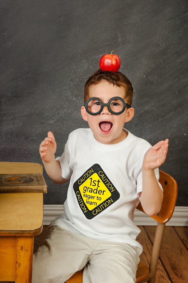 Może przedstawiać: Mały chłopiec w białej koszulce z ż&oacute;łtym znakiem ostrzegawczym, na kt&oacute;rym widnieje napis "1st grader eager to learn", siedzi na drewnianym krześle. Nosi duże, okrągłe, czarne okulary i ma na głowie czerwone jabłko.