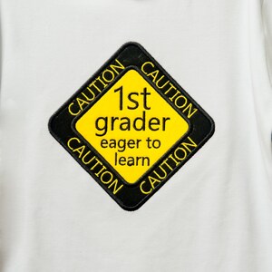 Może przedstawiać: Biała koszulka z ż&oacute;łto-czarnym znakiem ostrzegawczym z napisem "1st grader eager to learn".