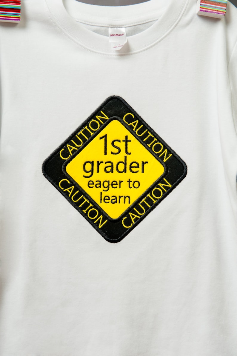 Może przedstawiać: Biała koszulka z ż&oacute;łto-czarnym znakiem ostrzegawczym z napisem "1st grader eager to learn".
