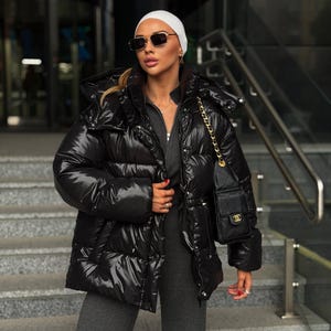 Puede incluir: Una chaqueta acolchada de color negro brillante con cuello alto, capucha y cierre de cremallera frontal. La chaqueta presenta paneles acolchados horizontales y mangas largas. Un bolso negro acolchado con cadena dorada se lleva al hombro.