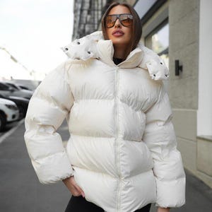 May include: A cream-colored puffer jacket featuring a high collar, a full-length front zipper, and a detachable hood with snap button closures. The jacket has a quilted, horizontal-baffled design with long sleeves and a slightly oversized fit.