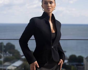 May include: A black blazer with a high collar and a single-button closure. The blazer is paired with a black dress. The background features a view of the ocean and a cityscape.