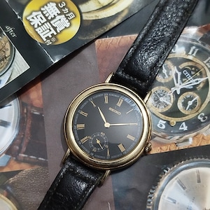 Op de afbeelding: Een vintage Seiko polshorloge met een zwarte wijzerplaat, goudkleurige lunette en zwarte leren band. Het horloge heeft gouden Romeinse cijfers en een kleine secondenwijzer. De achtergrond bevat horloge-gerelateerde afbeeldingen.