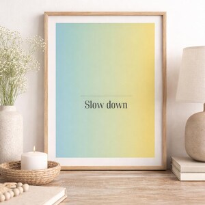 Könnte beinhalten: Ein gerahmter Druck mit einem Farbverlauf aus Blau, Grün und Gelb. Der Text "Slow down" steht unter einer dünnen Linie. Der Druck befindet sich auf einer Holzoberfläche mit einer Kerze, einer Vase und einer Lampe.