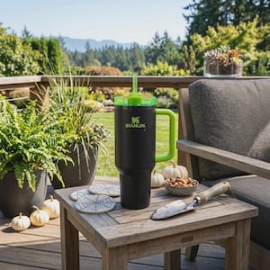 May include: A black Stanley tumbler with a green handle, lid, and straw sits on a wooden table. The tumbler has the Stanley logo in green. The table also holds spiderweb coasters, a small bowl of nuts, a trowel, and small pumpkins.
