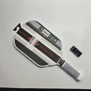 May include: A new JOOLA PRO PERSUES IV 16 mm pickleball paddle with a black and white design. The paddle has a gray handle with the text "TAP HERE" and a black grip tape.