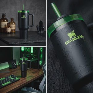 May include: Black Stanley tumbler with a green lid and straw. The tumbler has a handle and the Stanley logo in green. The design includes a spiderweb pattern and a green accent. The tumbler is shown in different settings, including a desk and a dark background.