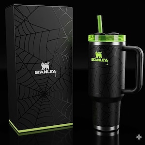 May include: Black Stanley tumbler with a green lid and straw, featuring a spiderweb design. The tumbler has a handle and the Stanley logo in green. A matching black box with a spiderweb design and the Stanley logo is also present.
