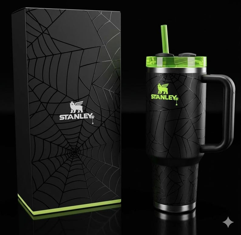 May include: Black Stanley tumbler with a green lid and straw, featuring a spiderweb design. The tumbler has a handle and the Stanley logo in green. A matching black box with a spiderweb design and the Stanley logo is also present.