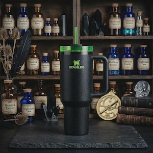 May include: Black Stanley tumbler with a green lid and straw, featuring a spiderweb design. The tumbler has a handle and the Stanley logo. The background includes apothecary bottles and a shelf.