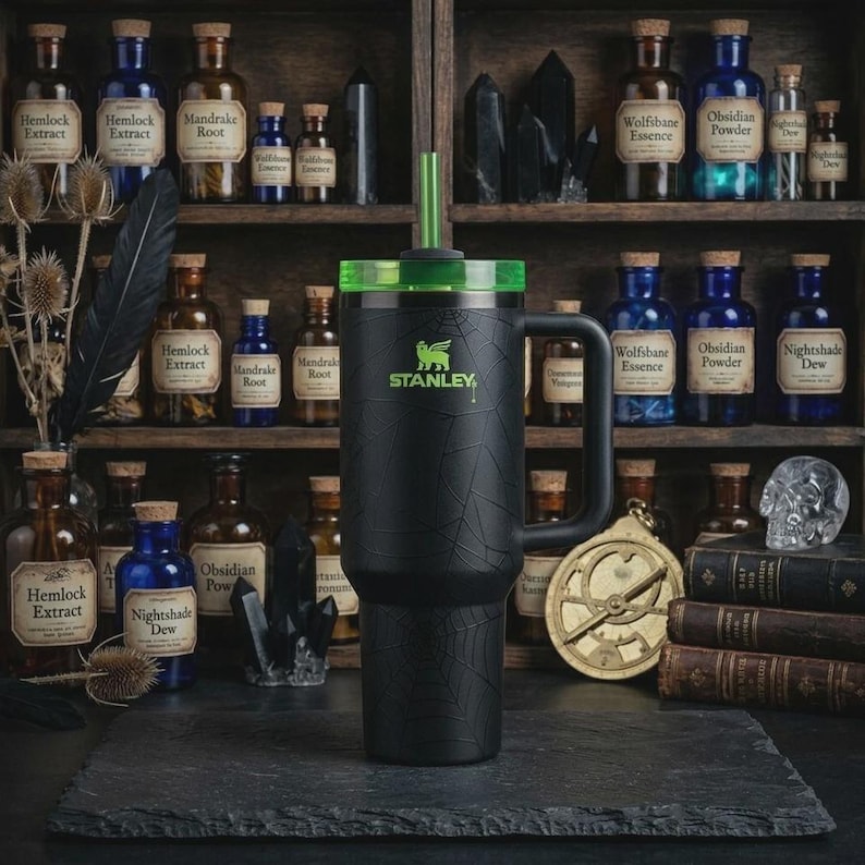 May include: Black Stanley tumbler with a green lid and straw, featuring a spiderweb design. The tumbler has a handle and the Stanley logo. The background includes apothecary bottles and a shelf.