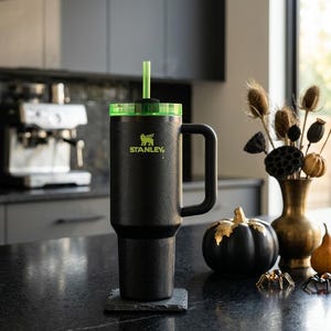 May include: Black Stanley tumbler with a green lid and straw, featuring the Stanley logo in neon green. The tumbler has a handle and a textured spiderweb design. It sits on a dark gray coaster, with a black pumpkin and decorative spiders nearby.
