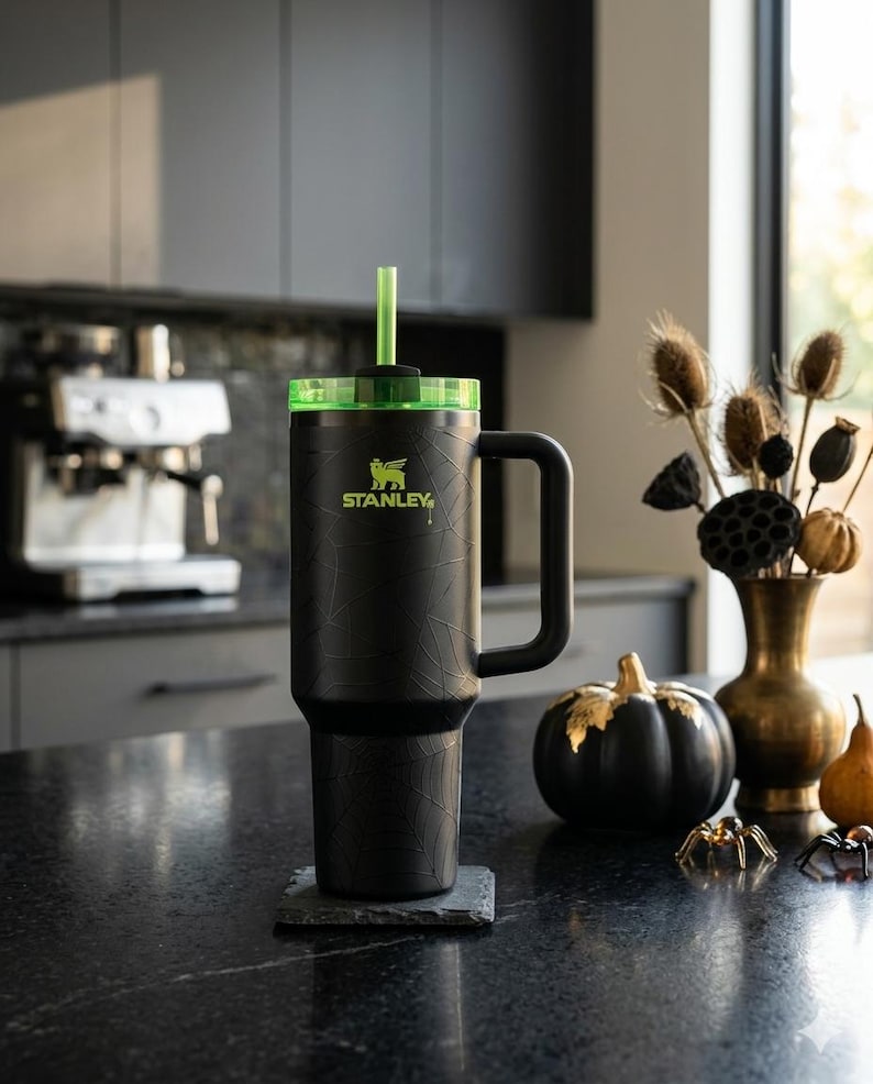 May include: Black Stanley tumbler with a green lid and straw, featuring the Stanley logo in neon green. The tumbler has a handle and a textured spiderweb design. It sits on a dark gray coaster, with a black pumpkin and decorative spiders nearby.
