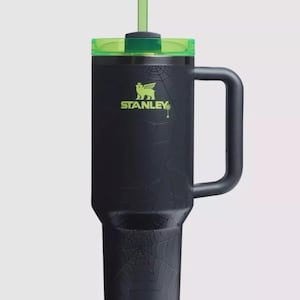 May include: Black Stanley tumbler with a green lid and straw. The tumbler features a spiderweb design and the Stanley logo in green. It has a large handle for easy carrying.