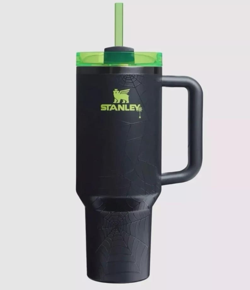 May include: Black Stanley tumbler with a green lid and straw. The tumbler features a spiderweb design and the Stanley logo in green. It has a large handle for easy carrying.