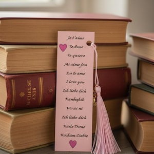 Peut inclure: Un marque-page rose avec un pompon, avec les mots "Je t'aime" et d'autres phrases exprimant l'amour dans différentes langues. Un cœur rose est en haut et en bas. Le marque-page est à côté d'une pile de livres.