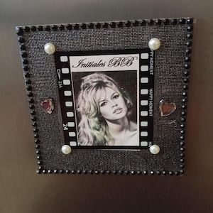 Op de afbeelding: Een rechthoekige, zwart omlijste magneet met een zwart-wit afbeelding van een vrouw. De lijst is versierd met kleine zwarte strass-steentjes en imitatieparels. De afbeelding heeft de tekst "Initiales BB".