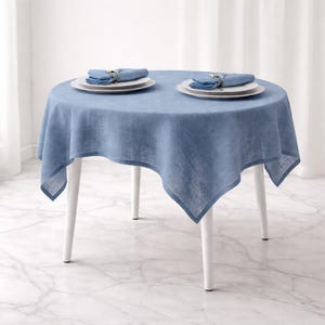 May include: A round table with a blue linen tablecloth, set with two plates, napkins, and gold napkin rings. The table has white legs and sits on a marble floor. The tablecloth drapes over the edges.