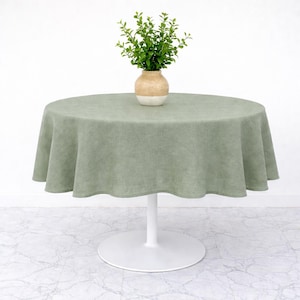 May include: A round, sage green linen tablecloth draped over a white pedestal table. A small vase with a beige and white finish holds a sprig of greenery, adding a touch of nature to the scene. The table is a stylish piece for dining.