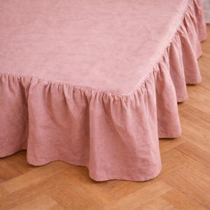 May include: A dusty rose bed skirt with a ruffled edge. The fabric appears to be linen or a similar material. The bed skirt is draped over a bed frame, with the ruffled edge hanging down. The background is a wooden floor.