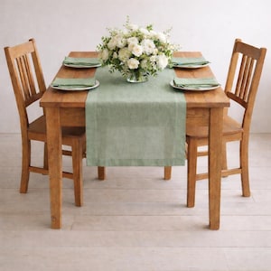 May include: A wooden dining table with two matching chairs. The table is set with a sage green table runner, white plates, and napkins. A vase of white flowers sits in the center. The table is approximately 122 cm long.