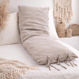 May include: A long, beige body pillow with tie closures at the end rests on a white bed. The pillow is made of a textured fabric and is surrounded by other white pillows. A woven wall hanging and decorative baskets are in the background.