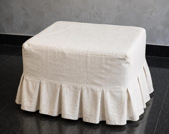 Puede incluir: Un otomano cuadrado tapizado en una tela de color blanco roto con un tejido texturizado. La base presenta una falda plisada que se extiende por los cuatro lados, creando un aspecto estructurado con pliegues verticales en el borde inferior.