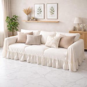 May include: A white sofa with a ruffled skirt, adorned with beige and striped throw pillows. The sofa is in a well-lit room with framed botanical prints and a wooden side table, creating a cozy living space.