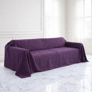 May include: A dark purple sofa cover draped over a three-seater sofa. The cover is made of a textured fabric and hangs over the arms of the sofa. The sofa is in a room with white walls and marble flooring.