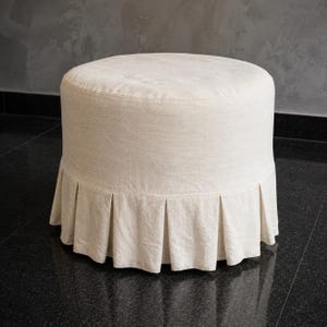 May include: A round, off-white ottoman featuring a textured fabric upholstery. The base of the ottoman is finished with a pleated skirt that extends around the entire circumference, providing a structured look to the cylindrical furniture piece.
