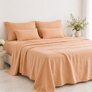 Pode incluir: Um conjunto de cama cor de pêssego, incluindo um lençol com elástico, um lençol de cima e quatro fronhas, é exibido em uma cama. A roupa de cama tem uma aparência texturizada. Um vaso de flores está em uma mesa próxima.