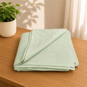 Op de afbeelding: Een stapel gevouwen, lichtgroene linnen servetten op een houten tafel. De servetten zijn netjes gerangschikt, met een hoek omgeslagen, waardoor de textuur van de stof zichtbaar is. Een kleine potplant staat op de achtergrond.