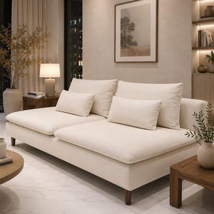 May include: A cream-colored sofa featuring two large back cushions and two rectangular throw pillows. The sofa is supported by short, dark wooden legs and has a low-profile design with a wide, deep seating area upholstered in a textured fabric.