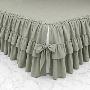 May include: A sage green bed skirt featuring a double-layered ruffled design along the edges. The fabric has a visible woven texture and includes a decorative bow tie detail at the corner, creating a layered appearance around the base of the bed.